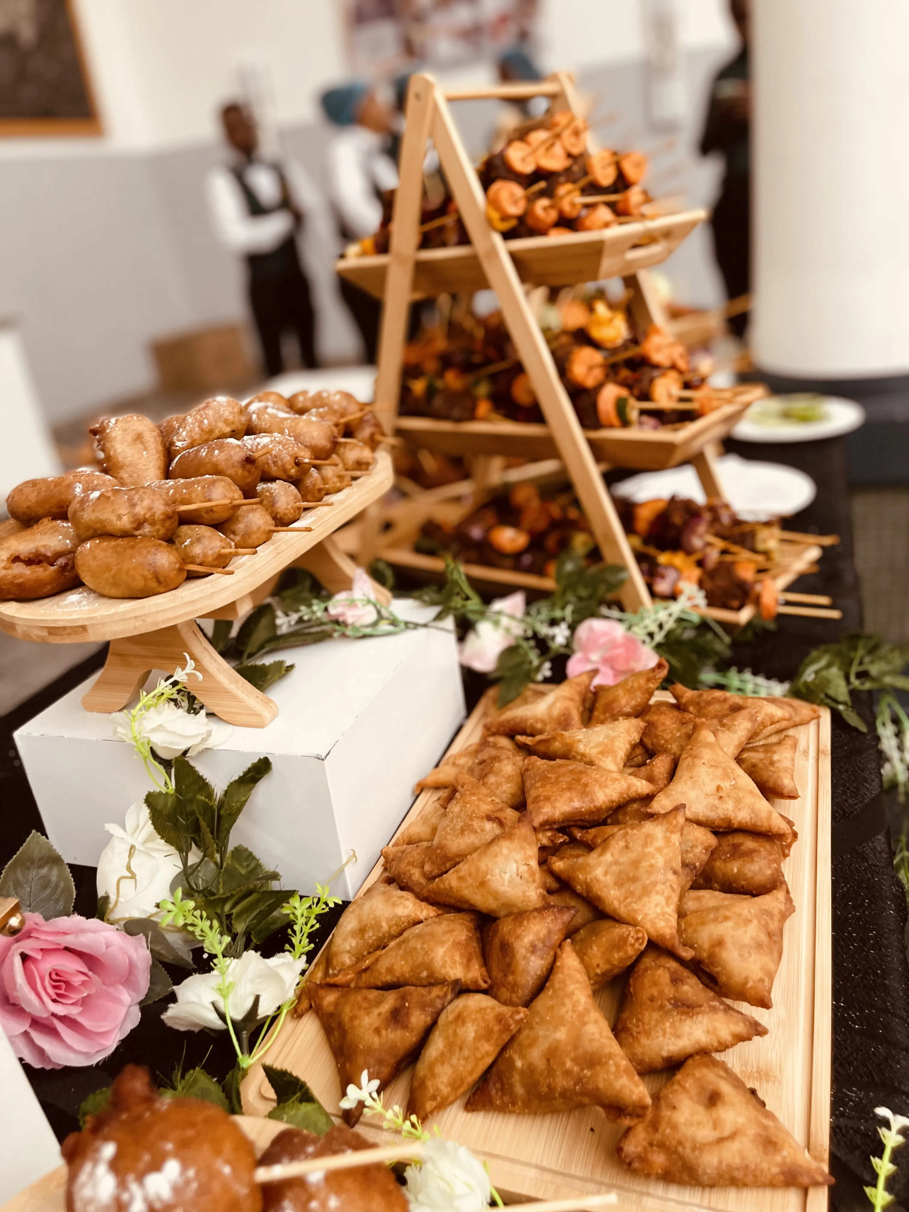 Elegant small chops display at an event with samosas, puff puff, and skewered meats on tiered wooden stands